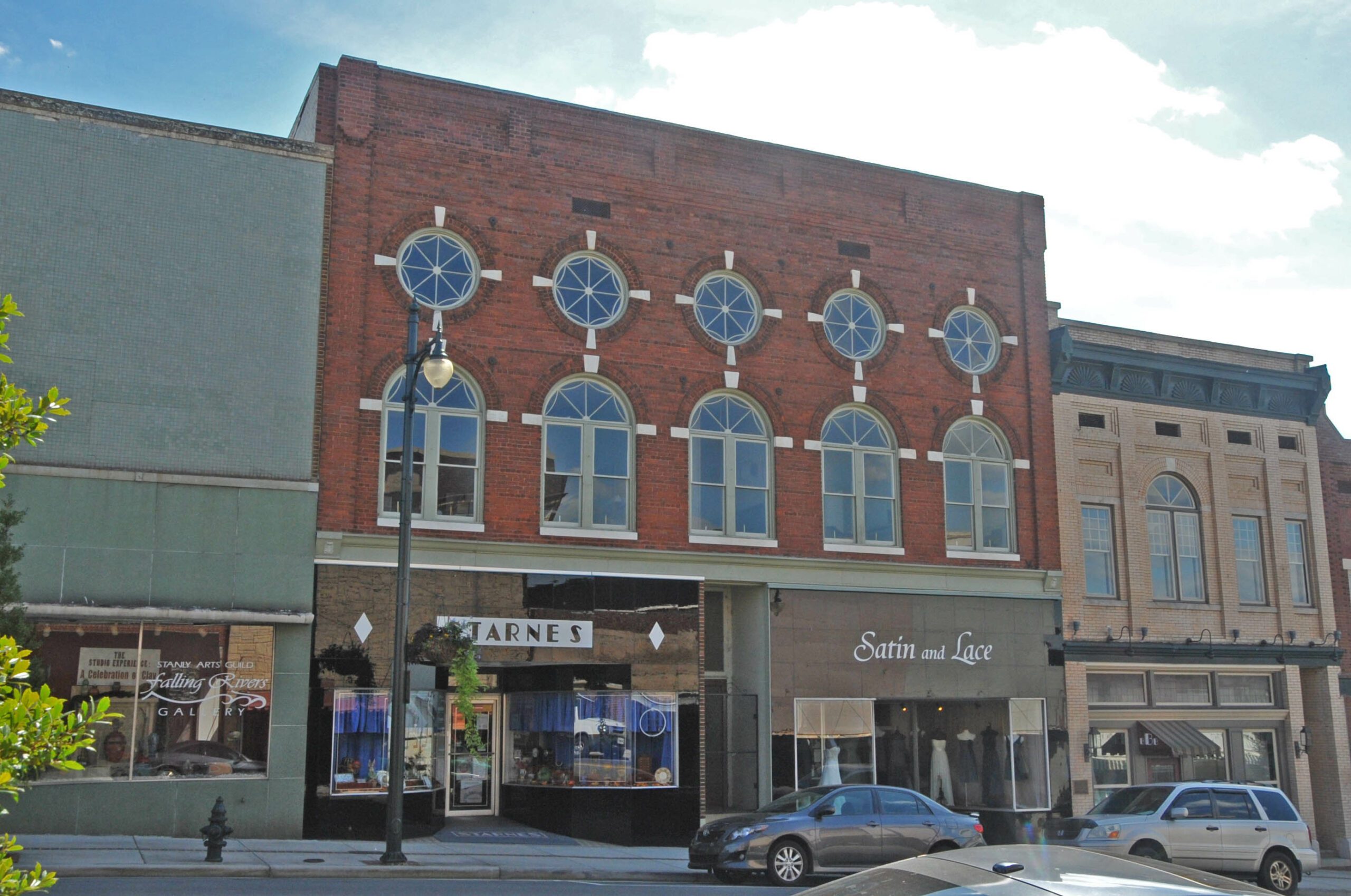 View of a Unique Red Brick Building With Stylish Round Window Frames, Housing Businesses Including ‘Tarnes’ and ‘Satin and Lace’ on a Sunny Day in a Downtown Street.